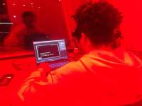 A person sits in front of a laptop computer. Their reflection also appears on a glass in front. The image has an orange hue.