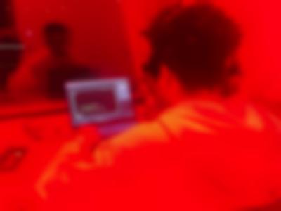 A person sits in front of a laptop computer. Their reflection also appears on a glass in front. The image has an orange hue.