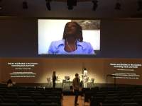 A group of people stand around the stage of an auditorium. Above them, an image of a person wearing earphones is being projected. Beneath it, to the right and to the left, two other projections show a slide with text, which reads 