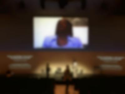 A group of people stand around the stage of an auditorium. Above them, an image of a person wearing earphones is being projected. Beneath it, to the right and to the left, two other projections show a slide with text, which reads 