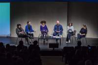 Six people sit on stage around a low table. The first rows of the audience are visible.
