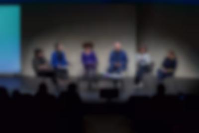 Six people sit on stage around a low table. The first rows of the audience are visible.