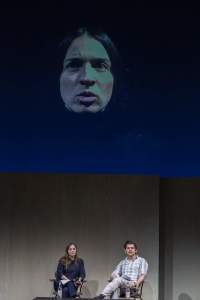 Two people sit on stage. Above them, the face of a person against a dark blue background is being projected.