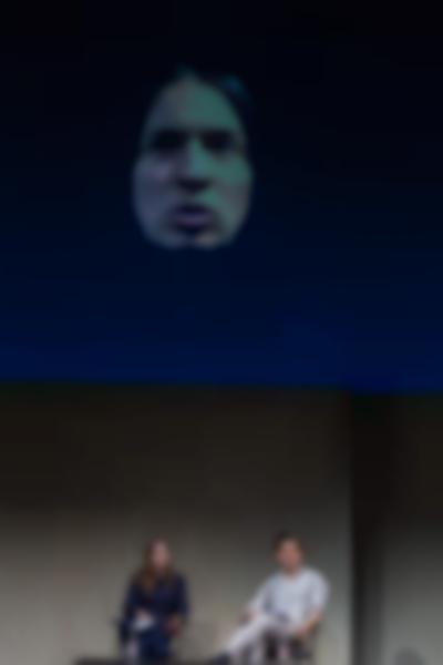 Two people sit on stage. Above them, the face of a person against a dark blue background is being projected.