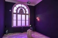 Indoor space with a large round top window and purple walls. In this space, there is an object on a plinth in the middle of the space, a neon sign on the wall, a mirror ball hanging from the ceiling and a small stool.