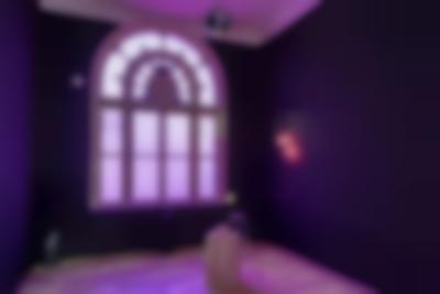 Indoor space with a large round top window and purple walls. In this space, there is an object on a plinth in the middle of the space, a neon sign on the wall, a mirror ball hanging from the ceiling and a small stool.
