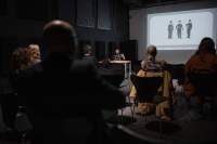 Several people, sitting on chairs, face the front of the room, where a person is delivering a presentation. Behind the presenter, a slide is being projected onto a screen.