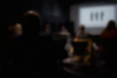 Several people, sitting on chairs, face the front of the room, where a person is delivering a presentation. Behind the presenter, a slide is being projected onto a screen.