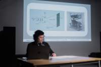 A person sits behind a desk and a computer, delivering a presentation. Behind them, a slide is being projected.