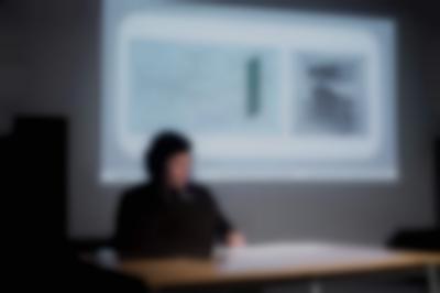 A person sits behind a desk and a computer, delivering a presentation. Behind them, a slide is being projected.