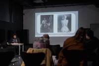 Several people, sitting on chairs, face the front of the room, where a person is delivering a presentation. Behind the presenter, a slide with two photographs is being projected onto a screen.