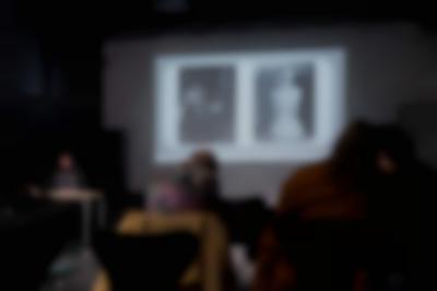 Several people, sitting on chairs, face the front of the room, where a person is delivering a presentation. Behind the presenter, a slide with two photographs is being projected onto a screen.