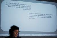 A person's head appears in front of a powerpoint slide projection. The white slide asks two questions about how the usage of photography and video throughout the twentieth century, which appear in black.