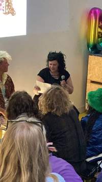 Two people sit in front of a group of people. The person on the right holds a sheet of paper and a microphone.