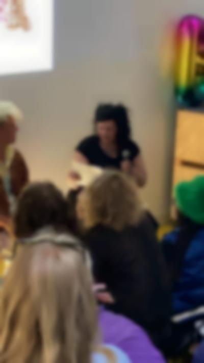 Two people sit in front of a group of people. The person on the right holds a sheet of paper and a microphone.