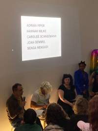 Three people sit in front of a group of people. The name of several artists (Adrian Piper, Hannah Wilke, Carolee Schneemann, Joan Semmel and Senga Nengudi) is being projected in black text above them. The person to the left is holding a microphone.