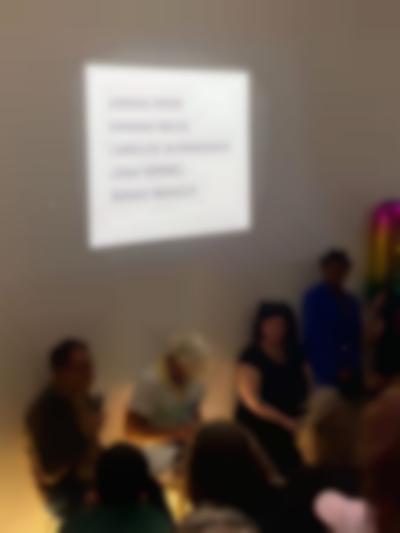 Three people sit in front of a group of people. The name of several artists (Adrian Piper, Hannah Wilke, Carolee Schneemann, Joan Semmel and Senga Nengudi) is being projected in black text above them. The person to the left is holding a microphone.