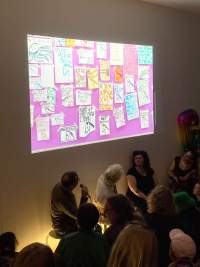 Three people sit in front of a group of people. Above them, an image of several drawings stuck to a pink surface is being projected. The two people to the left look up to the image.