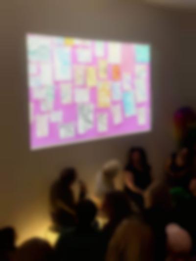 Three people sit in front of a group of people. Above them, an image of several drawings stuck to a pink surface is being projected. The two people to the left look up to the image.