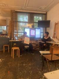 Two people are having a discussion in front of a screen showing an image. One of them is holding a poster, which the other person looks towards.