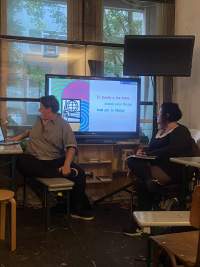 Two people are having a discussion in front of a screen showing an image and some text. One of them is looking at the screen and the other faces their laptop.