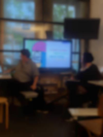 Two people are having a discussion in front of a screen showing an image and some text. One of them is looking at the screen and the other faces their laptop.