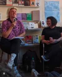 Two people sit next to each other. The person on the left holds a microphone. The person on the right is laughing and looking at the other person. There are bookshelves behind them.
