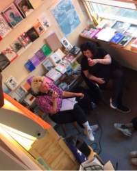 Seen from above, two people sit next to each other. The person on the left holds a microphone. The person on the right looks at them, while taking a sip from a drink. There are bookshelves behind them.