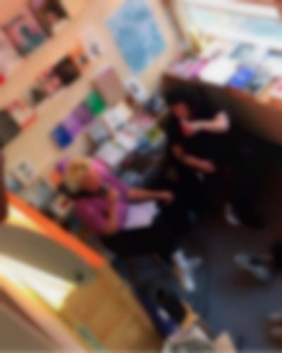 Seen from above, two people sit next to each other. The person on the left holds a microphone. The person on the right looks at them, while taking a sip from a drink. There are bookshelves behind them.