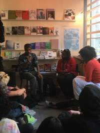 Two people sit in front of a group of people. The person on the left holds a microphone. There are bookshelves behind them.