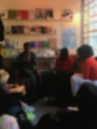 Two people sit in front of a group of people. The person on the left holds a microphone. There are bookshelves behind them.