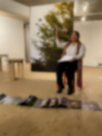 A person sits in front of a large photographic print that hangs from the ceiling in the middle of a space. The print partially depicts green foliage. They hold a piece of paper and are gesturing with their right hand. In front, a long line of flower prints stretches across the floor.