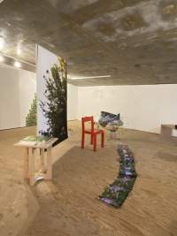 In an exhibition space, a large photographic print with green foliage is suspended from the ceiling. To its sides, we can find a red chair (right) and a wooden table with some photographic material on top (left). To its front, a long line of flower prints stretches across the floor.
