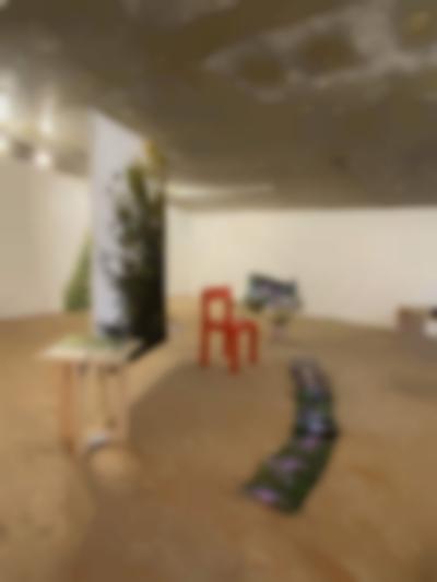 In an exhibition space, a large photographic print with green foliage is suspended from the ceiling. To its sides, we can find a red chair (right) and a wooden table with some photographic material on top (left). To its front, a long line of flower prints stretches across the floor.