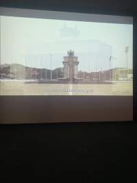 A film is being projected onto a screen. The captured frame shows a monument with a star on top. Behind it, several flag poles remain empty. The subtitle reads: 