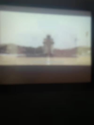 A film is being projected onto a screen. The captured frame shows a monument with a star on top. Behind it, several flag poles remain empty. The subtitle reads: 
