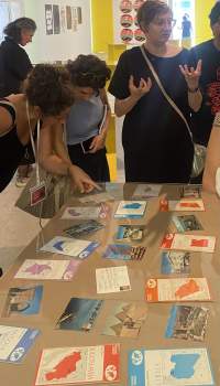 Travel postcards and brochures have been set on a table. A group of people is standing around it. Two people observe one of the postcards closely.