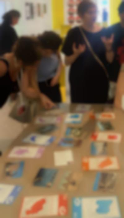 Travel postcards and brochures have been set on a table. A group of people is standing around it. Two people observe one of the postcards closely.