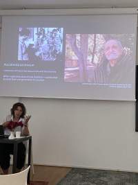 A person sits at a table delivering a presentation. Behind them a slide is being projected. The slide is titled 'Accidental archivism' and shows two images. One of a group of people around a camera and another of a male presenting person in a natural setting.