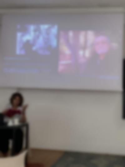 A person sits at a table delivering a presentation. Behind them a slide is being projected. The slide is titled 'Accidental archivism' and shows two images. One of a group of people around a camera and another of a male presenting person in a natural setting.