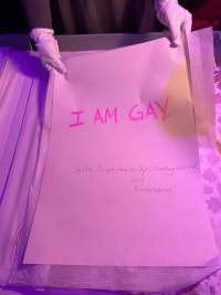 Two hands, wearing white gloves, hold a large white sheet of paper, which reads 