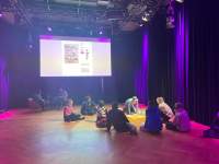 A group of people sit cross-legged on the floor. They are facing another person, who is on the spotlight. Behind them, a webpage is being projected on a large screen.