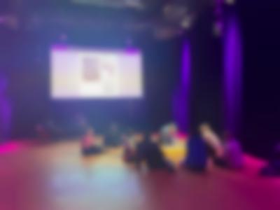 A group of people sit cross-legged on the floor. They are facing another person, who is on the spotlight. Behind them, a webpage is being projected on a large screen.