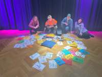 Four people pose behind several colourful sheets of paper with drawings, which are arranged in a circle on the floor. In the middle of the drawings, there is a pillow.