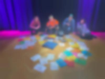 Four people pose behind several colourful sheets of paper with drawings, which are arranged in a circle on the floor. In the middle of the drawings, there is a pillow.