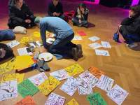 A person paints a sheet of paper on the floor. Around them, there are several other drawings. A group of people sit crossed-legged around the painter and the drawings, watching.