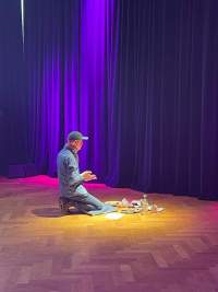 A person sits on their heels in front of several art supplies. They face the curtains, to the back of the space.