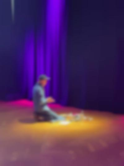 A person sits on their heels in front of several art supplies. They face the curtains, to the back of the space.