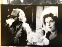 A feminine-presenting person holds a baby and faces the camera. To the left, a masculine-presenting person, wearing sunglasses, looks away.