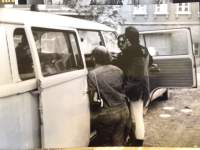 A group of three masculine-presenting people stand by the open door of a small Volkswagen bus, facing the inside.